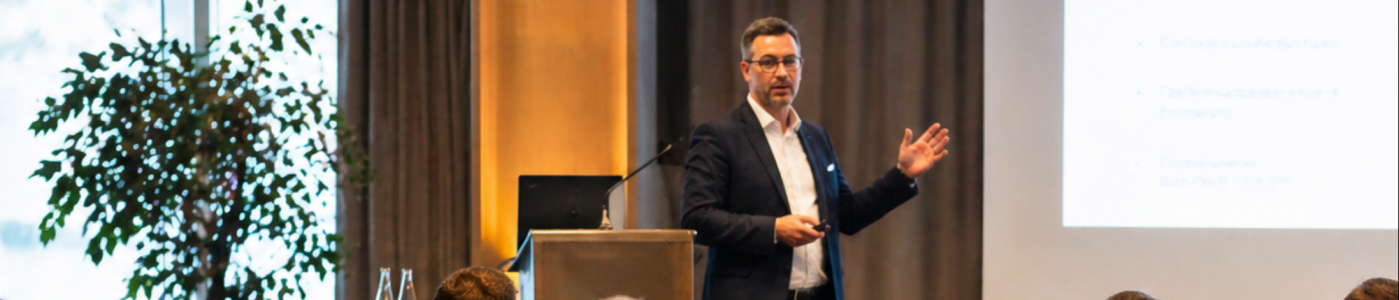 Conférencier en costume présentant devant un public assis dans une salle de réunion.