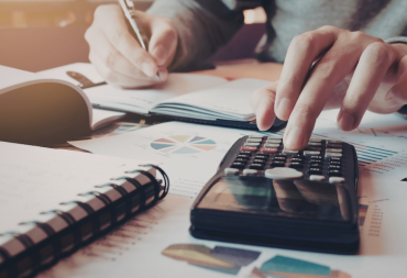 Personne utilisant une calculatrice et prenant des notes sur des documents financiers étalés sur un bureau.